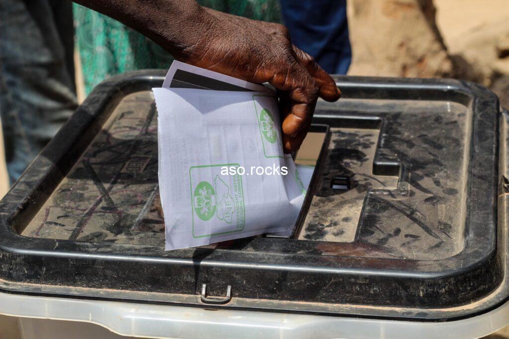 A hand casting a paper ballot into a Nigerian election voting box at a polling station